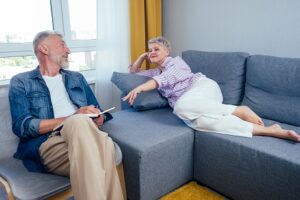 An older man and woman are conversing in a living room. The man sits with a notebook while the woman lies on a gray sofa, gesturing with her hand.